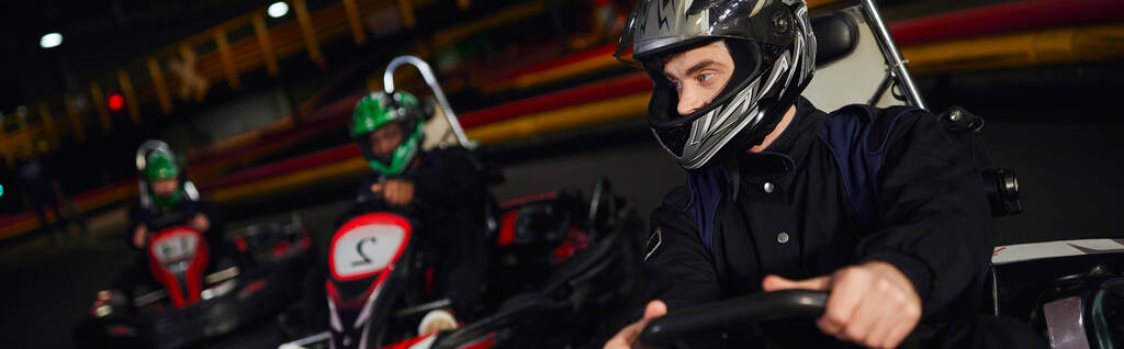 geconcentreerde man rijden go kart in de buurt van diverse bestuurders in helmen op indoor circuit, banner - Foto, afbeelding