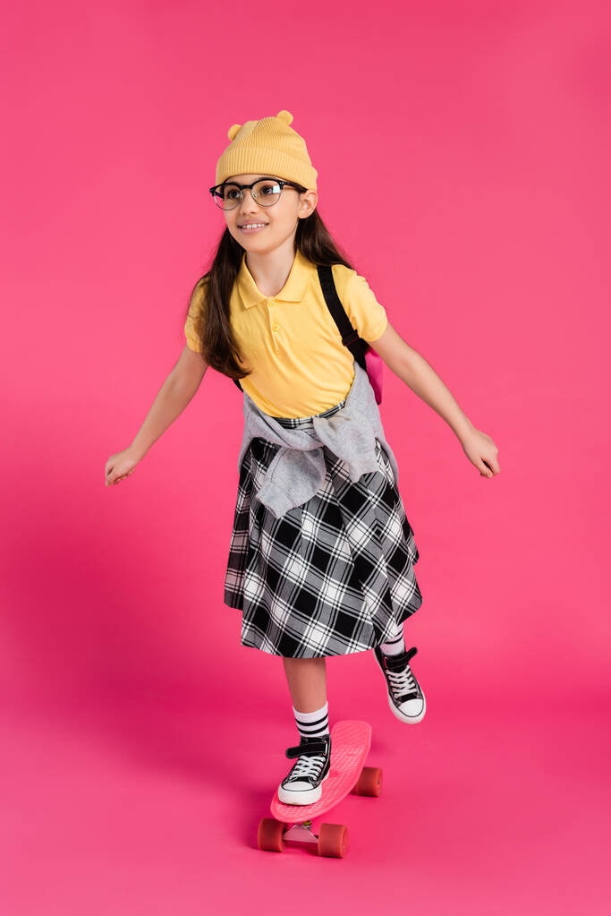 alegre colegiala en gorro sombrero y gafas de montar penny board sobre fondo rosa, aspecto elegante - Foto, imagen