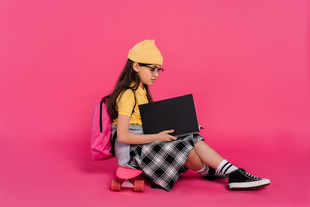 stylish schoolgirl in beanie hat and glasses sitting on penny board, pink background, notebooks - Photo, Image
