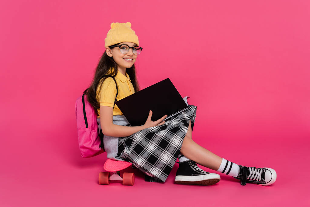 ペニーボードに座っているビーニー帽子とメガネの幸せな女子高生,ピンクの背景,ノートブック - 写真・画像