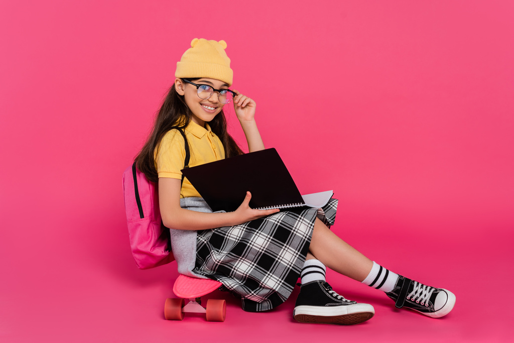 vrolijk schoolmeisje in muts en bril zitten op penny board, roze achtergrond, notitieboekjes - Foto, afbeelding