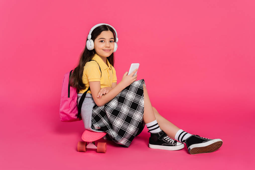 Kulaklıklı mutlu kız öğrenci, tahta üzerinde oturuyor, pembe arka plan, akıllı telefon, dijital... - Fotoğraf, Görsel