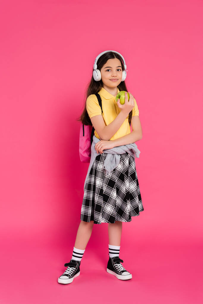 mosolygós iskolás lány vezeték nélküli fejhallgató gazdaság zöld alma rózsaszín háttér, gyerek hátizsák - Fotó, kép