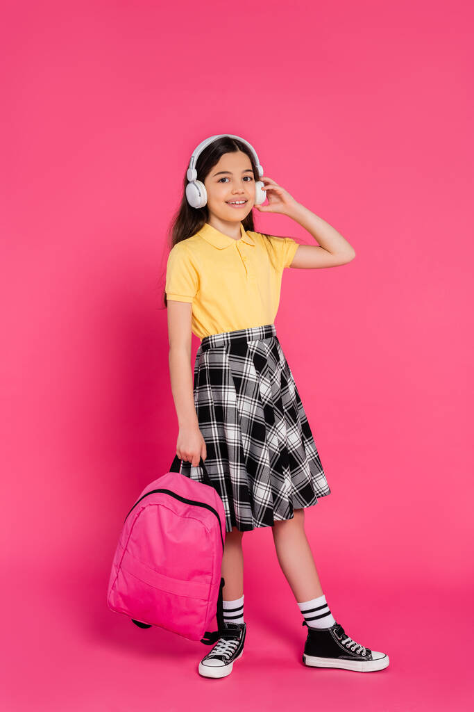 excited schoolgirl in wireless headphones holding backpack on pink background, brunette student - Photo, Image