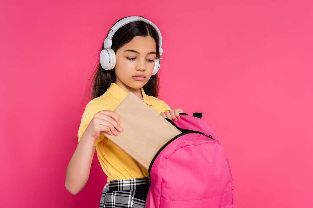 brunette schoolmeisje in draadloze hoofdtelefoon zetten boek in van rugzak, roze achtergrond, student - Foto, afbeelding