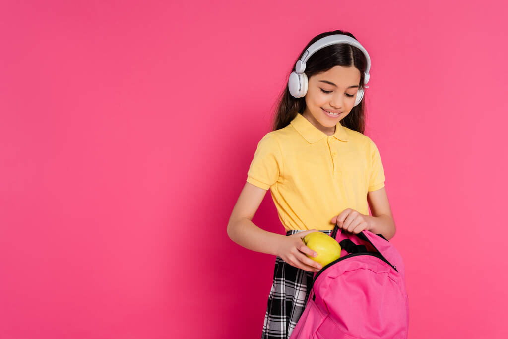 happy schoolgirl in wireless headphones putting apple inside of backpack, pink background, student - Photo, Image