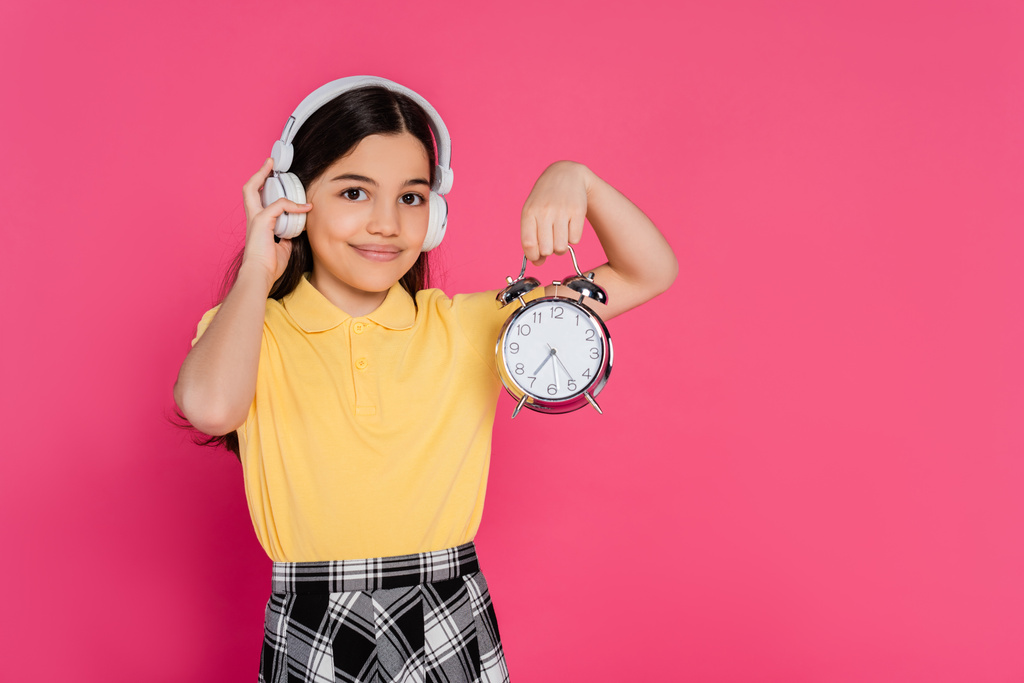 ピンクで隔離されたヴィンテージの目覚まし時計を保持するワイヤレスヘッドフォンの幸せな女子生徒,学生生活 - 写真・画像