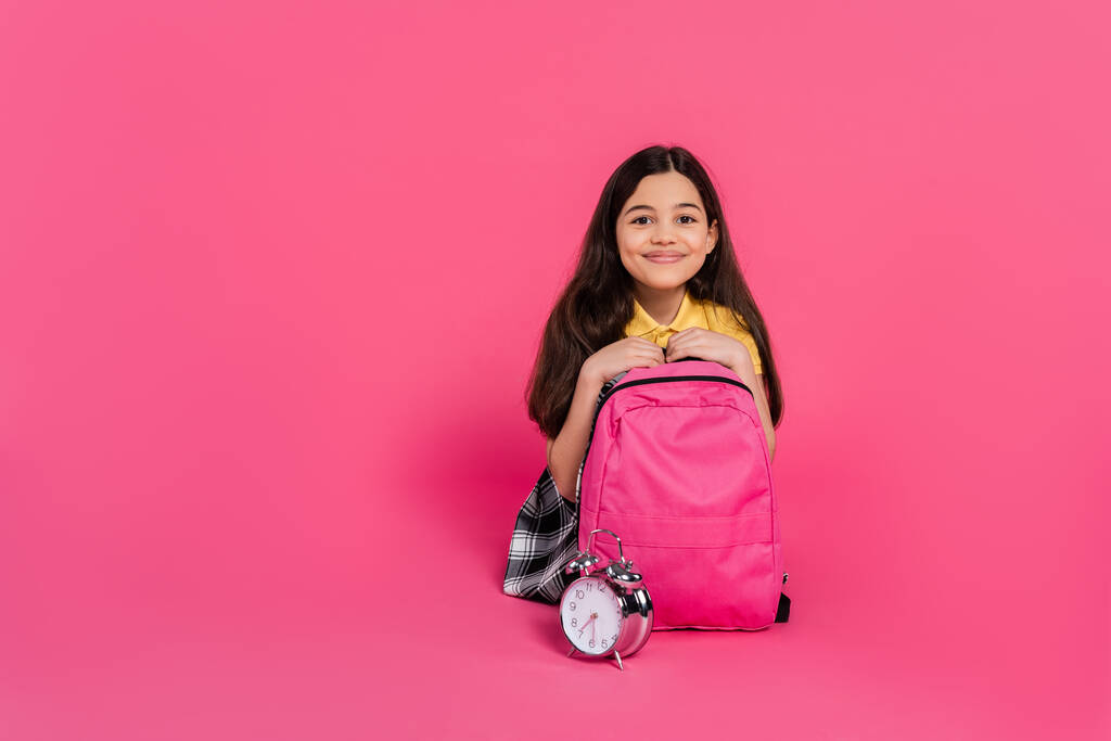 alegre morena estudante sentado com mochila perto de despertador vintage no fundo rosa - Foto, Imagem