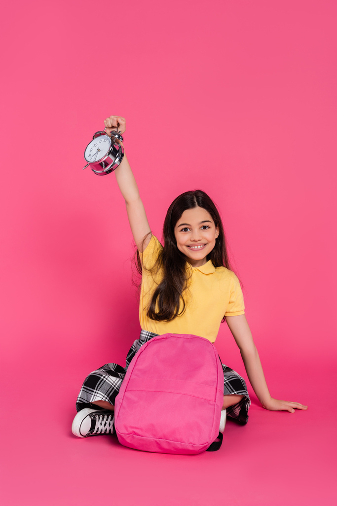 positive and brunette schoolgirl sitting with backpack, holding vintage alarm clock on pink - Photo, Image