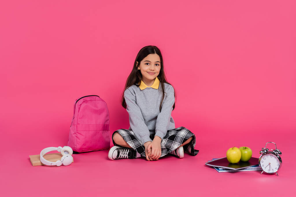 happy schoolgirl sitting near backpack, notebooks, headphones, green apples, vintage alarm clock - Photo, Image