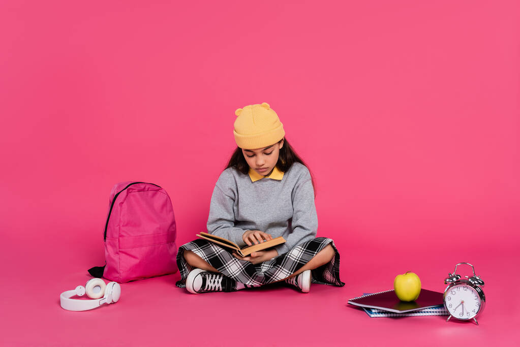 школярка в капелюсі Беані читає книгу, сидячи біля навушників, яблуко, рюкзак, будильник - Фото, зображення