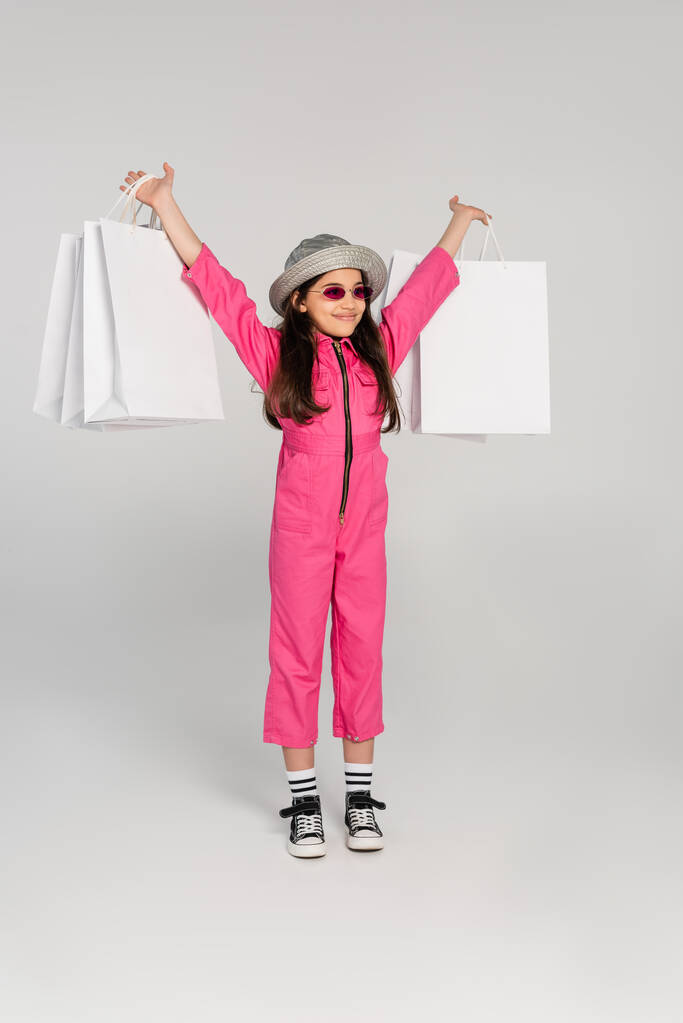 eccitata ragazza in elegante abito rosa e cappello panama che tiene borse della spesa su sfondo grigio - Foto, immagini