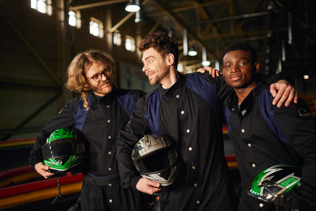 Gruppe multikultureller und fröhlicher Kartfahrer in Schutzanzügen, die sich umarmen und Helme tragen - Foto, Bild