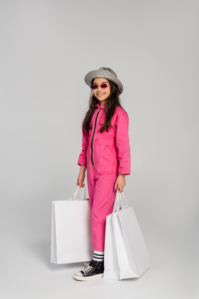 chica alegre en traje elegante, gafas de sol y sombrero panama sosteniendo bolsas de compras sobre fondo gris - Foto, imagen