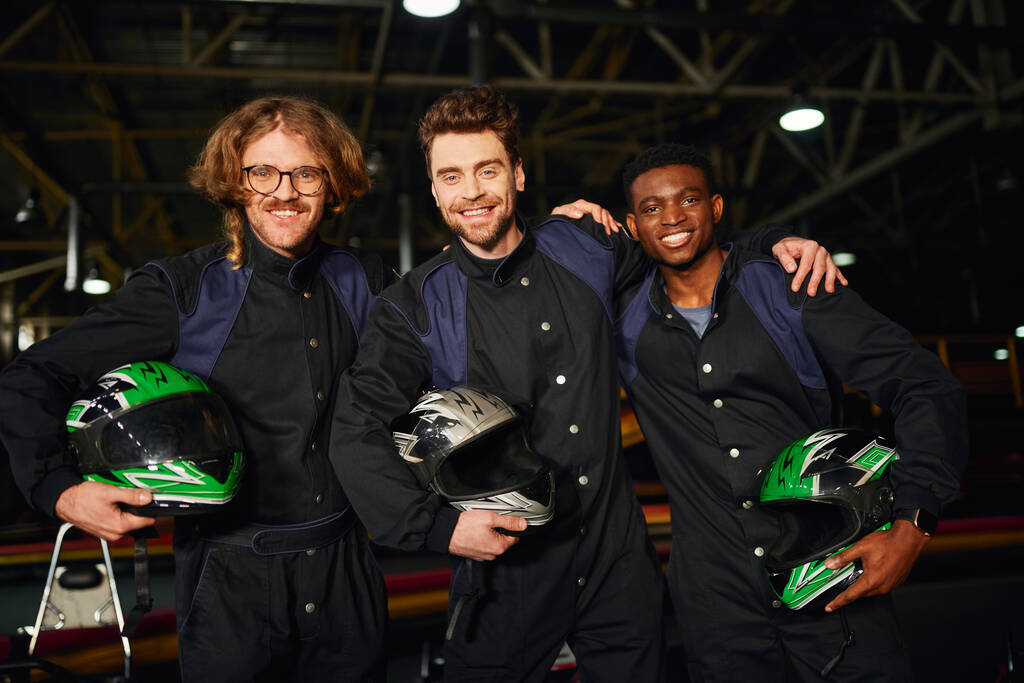 Gruppe multikultureller und fröhlicher Kartfahrer in Schutzanzügen, die sich umarmen und Helme tragen - Foto, Bild