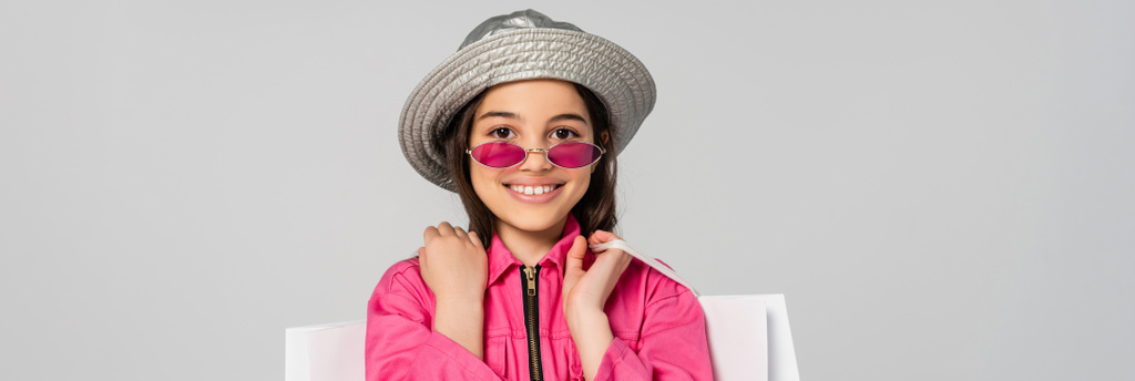 pozitivní dívka ve stylovém oblečení, slunečních brýlích a panamském klobouku pózující s nákupními taškami na šedé, banner - Fotografie, Obrázek