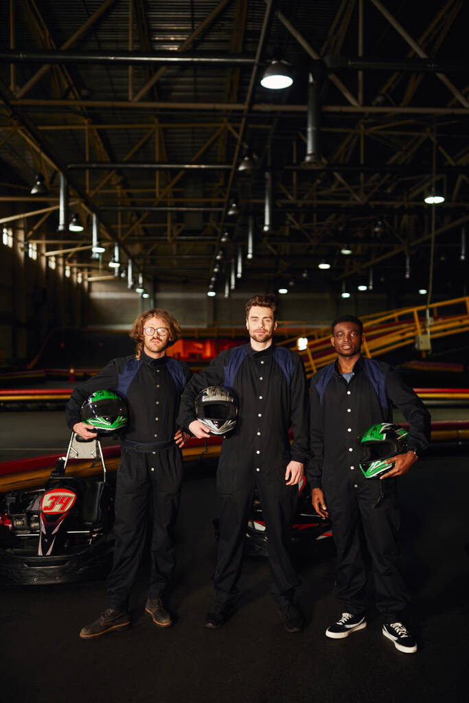 multicultural go kart drivers in protective suits standing and holding helmets, competitors - Photo, Image