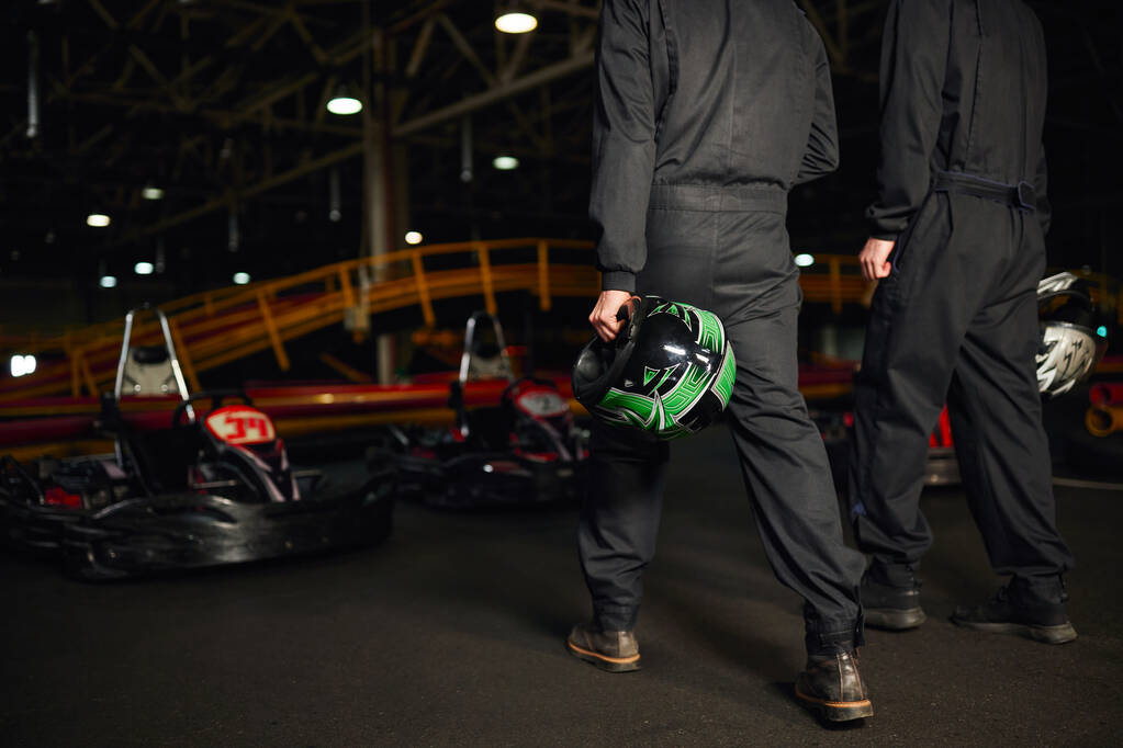 vista recortada de los conductores de karts go en ropa deportiva caminando en el circuito y la celebración de cascos, competidores - Foto, Imagen