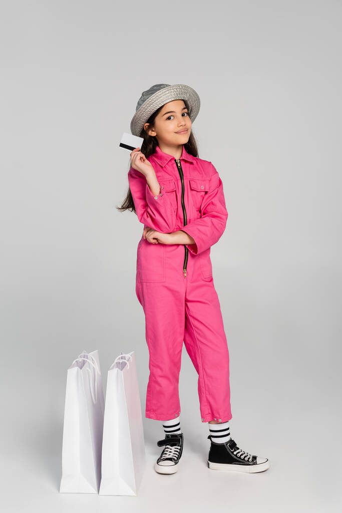 щаслива дівчина в модному вбранні і панама капелюх позує біля сумки для покупок, тримаючи кредитну картку на сірому - Фото, зображення