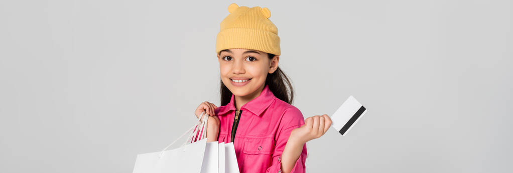 glückliches Mädchen mit Mütze, Kreditkarte isoliert auf grauem, trendigem Outfit, Kindereinkauf, Banner - Foto, Bild