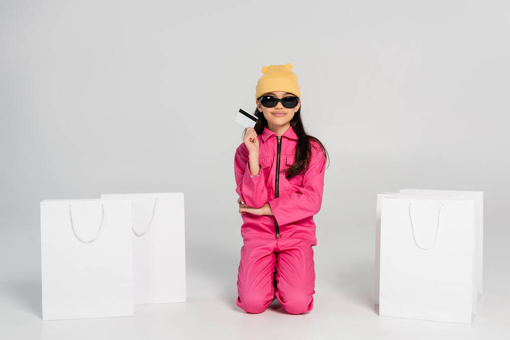 Şapkalı ve güneş gözlüklü mutlu bir kız gri, çocuk alışverişinde, moda kıyafetinde kredi kartı tutuyor. - Fotoğraf, Görsel