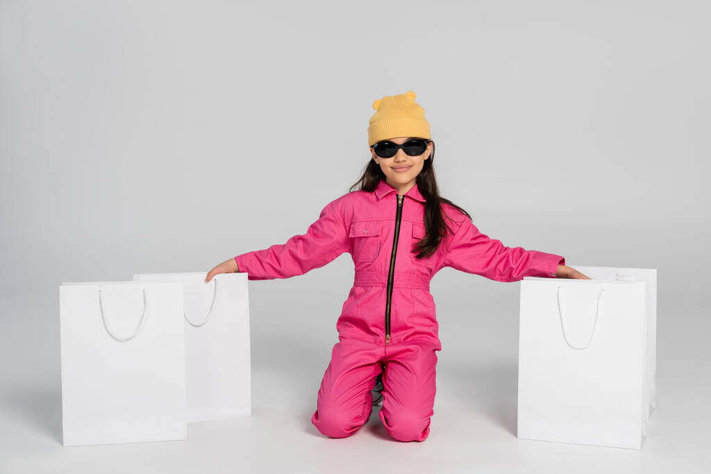 Bereli ve güneş gözlüklü neşeli kız gri alışveriş çantalarının yanında oturuyor. - Fotoğraf, Görsel