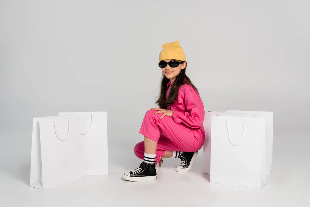 фешенебельная девушка в шапочке и солнцезащитных очках, сидящая рядом с сумками для покупок на сером, счастливом ребенке - Фото, изображение