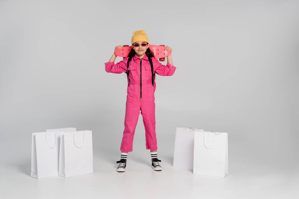 šťastná dívka v beanie a stylové sluneční brýle držení penny palubě, nákupní tašky na šedém pozadí - Fotografie, Obrázek