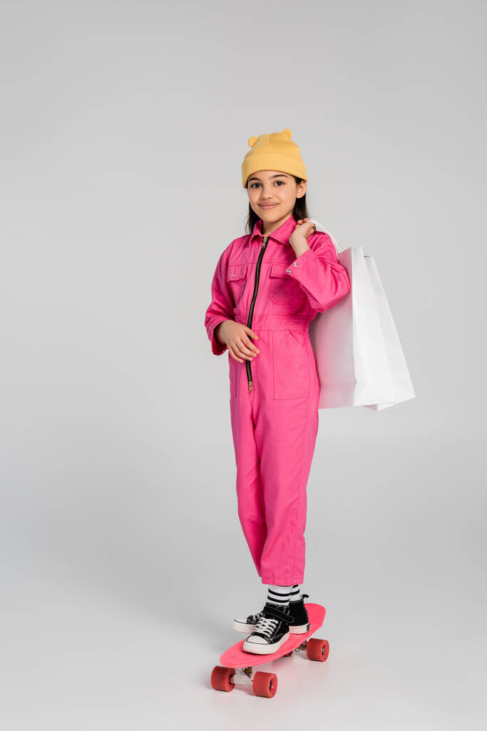 šťastná dívka v čepici a růžové oblečení na koni penny palubě a držení nákupních tašek na šedé - Fotografie, Obrázek