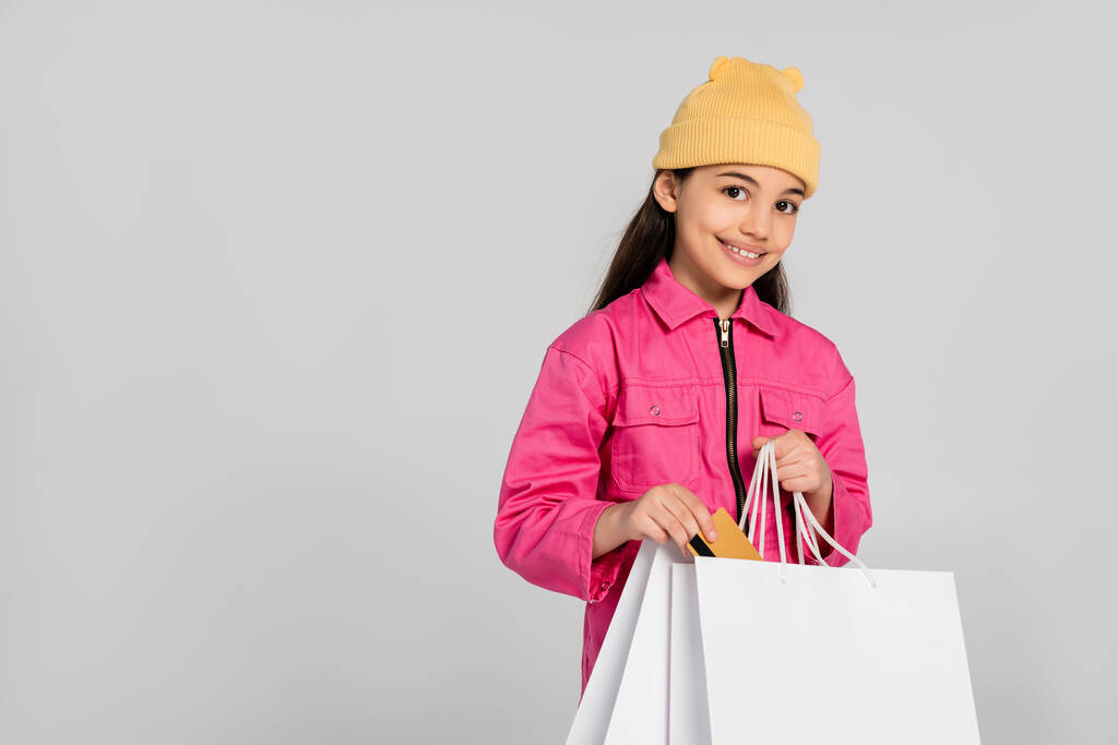 Bereli mutlu kız gri kıyafetler içinde kredi kartını alışveriş çantasına koyuyor, çocuk alışverişi yapıyor. - Fotoğraf, Görsel