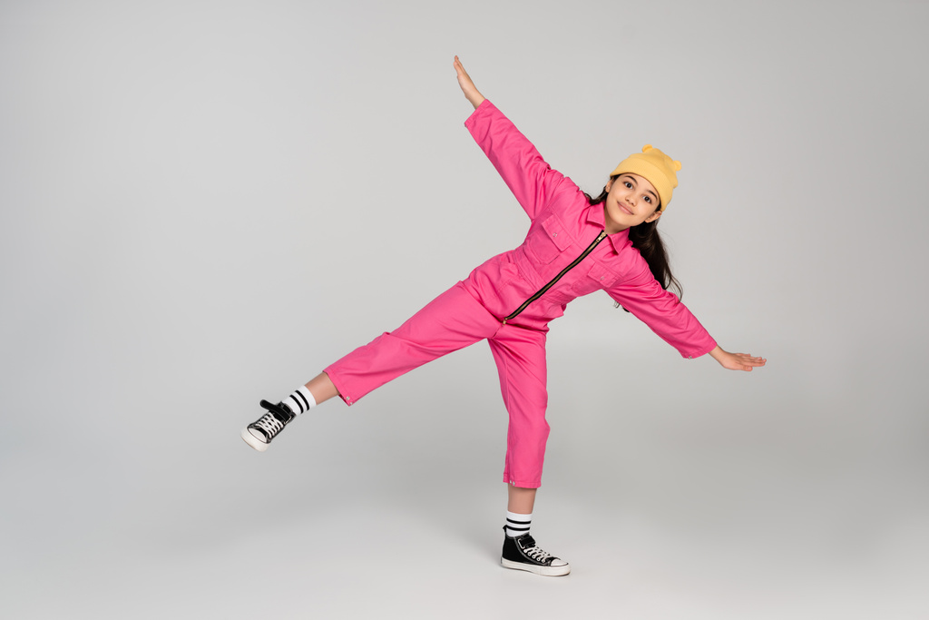 счастливая девушка в шляпе и стильном розовом наряде, позирующая с протянутыми руками на сером, веселится - Фото, изображение