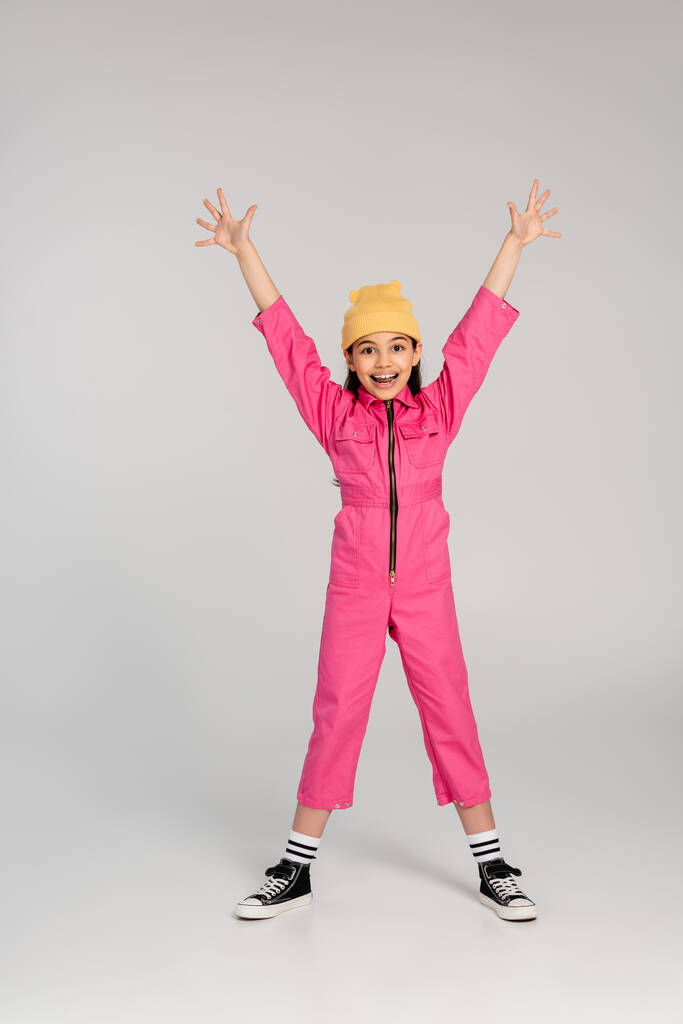 happy girl in beanie hat and pink outfit standing with outstretched hands on grey, have fun, style - Photo, Image