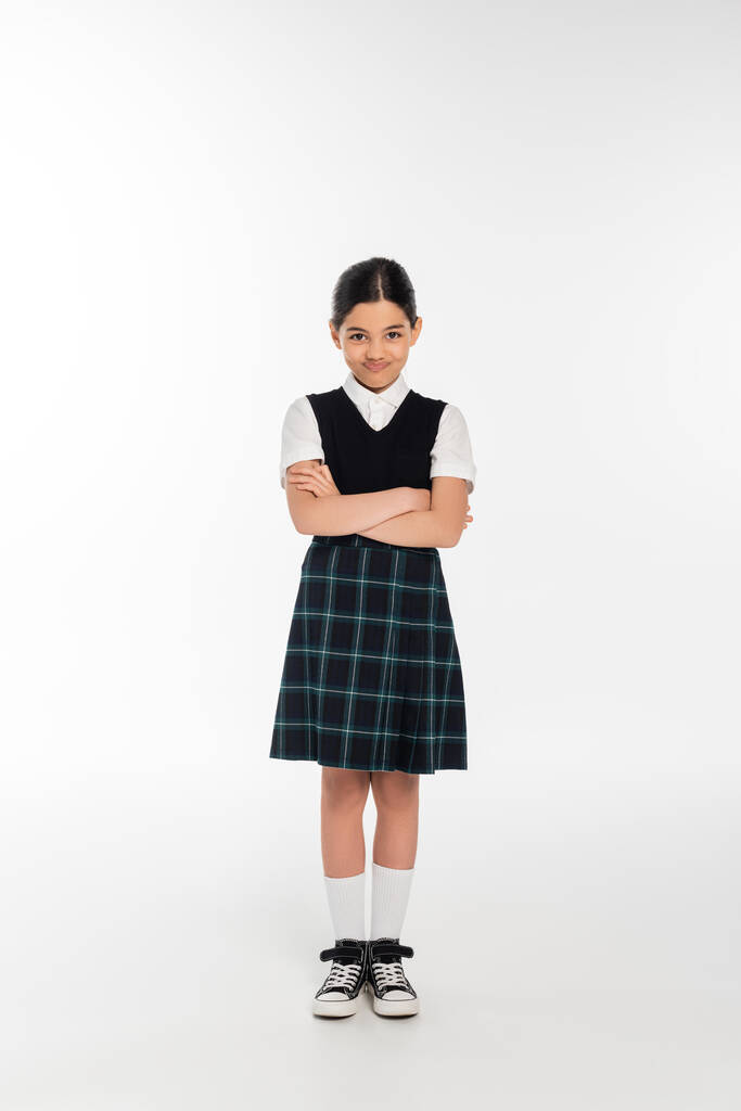 full length, offended schoolkid in uniform looking at camera and standing with folded arms on white - Photo, Image