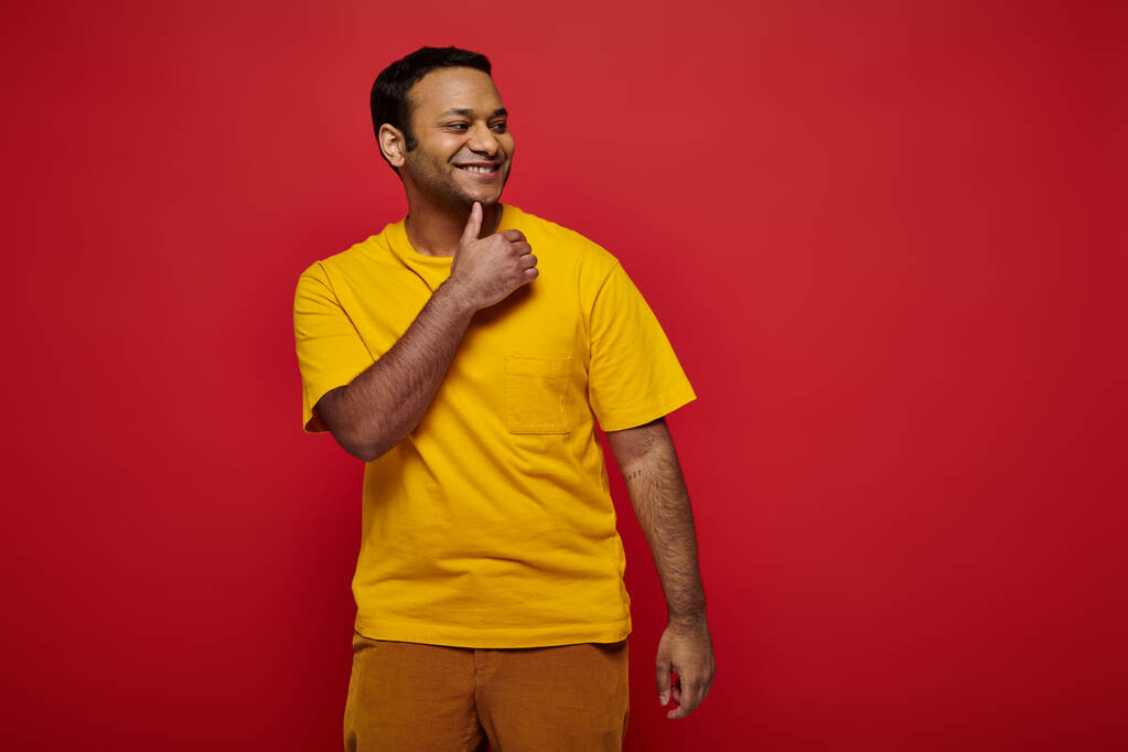 felice uomo indiano in abiti casual luminosi in piedi e sorridente su sfondo rosso in studio - Foto, immagini