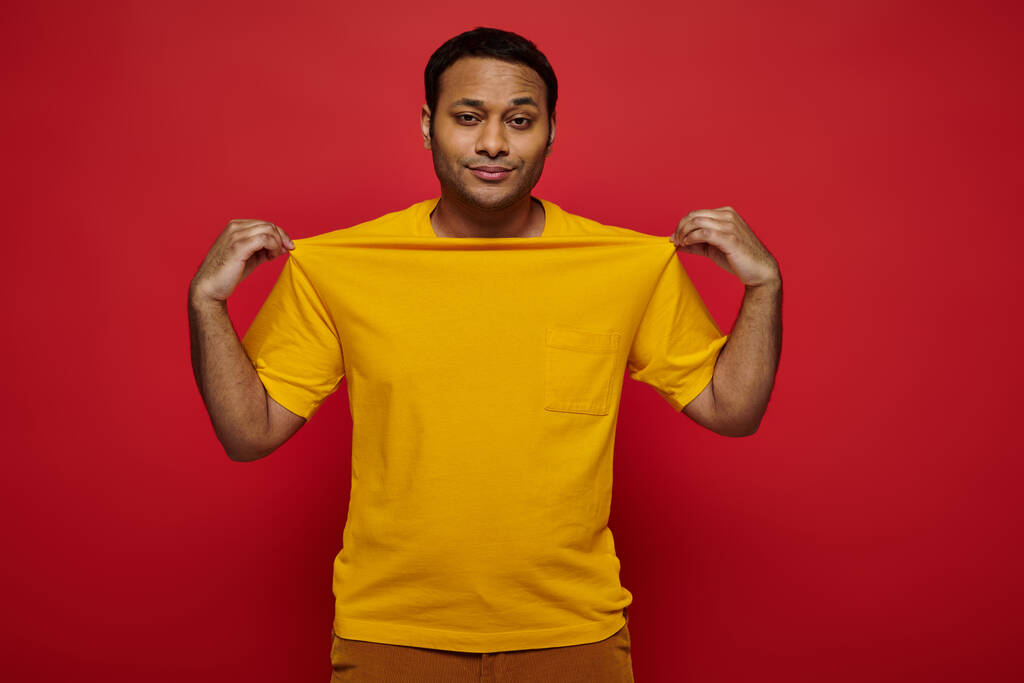 homme indien à la mode touchant t-shirt jaune et regardant la caméra sur fond rouge, style personnel - Photo, image