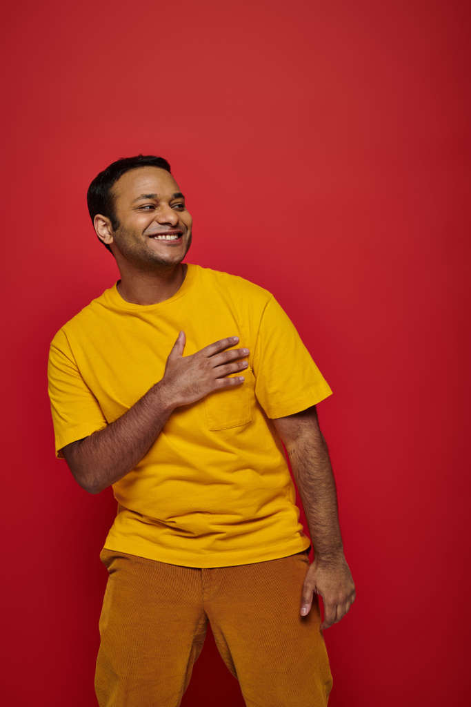 grateful indian man in bright casual clothes smiling and looking away on red background in studio - Photo, Image