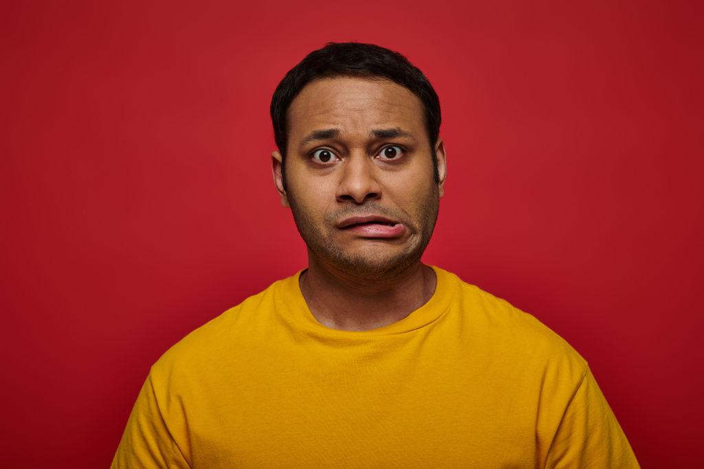 worried indian man in bright clothes looking at camera and grimacing on red background, stroke - Photo, Image