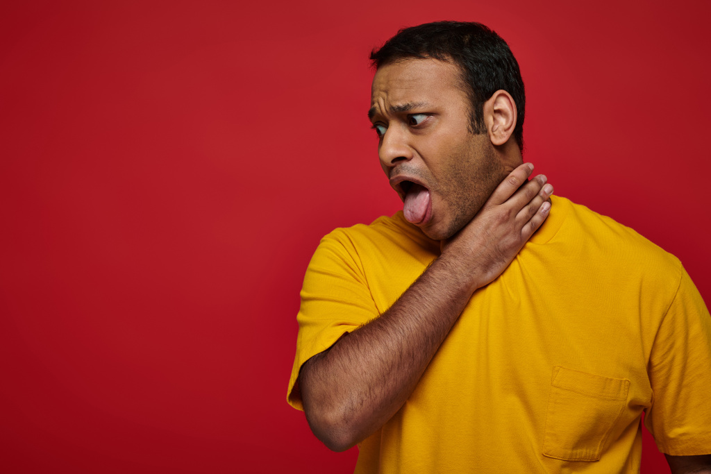 Ινδός άνδρας με κίτρινα ρούχα προεξέχει γλώσσα, ενώ πνίγεται στο κόκκινο φόντο, δυσκολία στην αναπνοή - Φωτογραφία, εικόνα