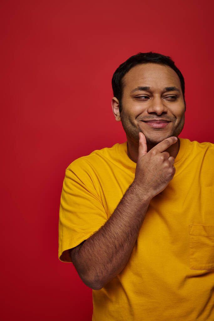 pleased indian man in yellow t-shirt touching chin and looking away on red background, thoughtful - Photo, Image