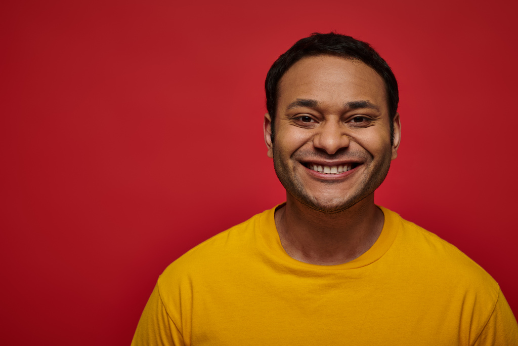 pozitív érzelem, boldog indiai férfi sárga pólóban mosolyog vagy vigyorog piros háttér stúdió - Fotó, kép