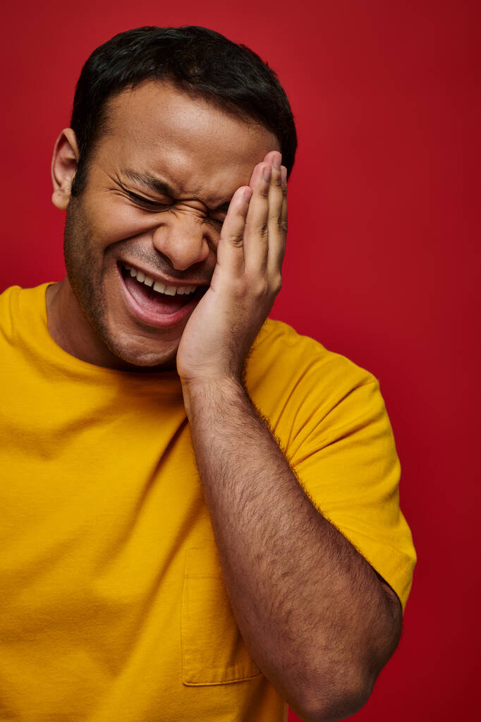 face expression, embarrassed indian man in yellow t-shirt laughing and touching face on red backdrop - Photo, Image
