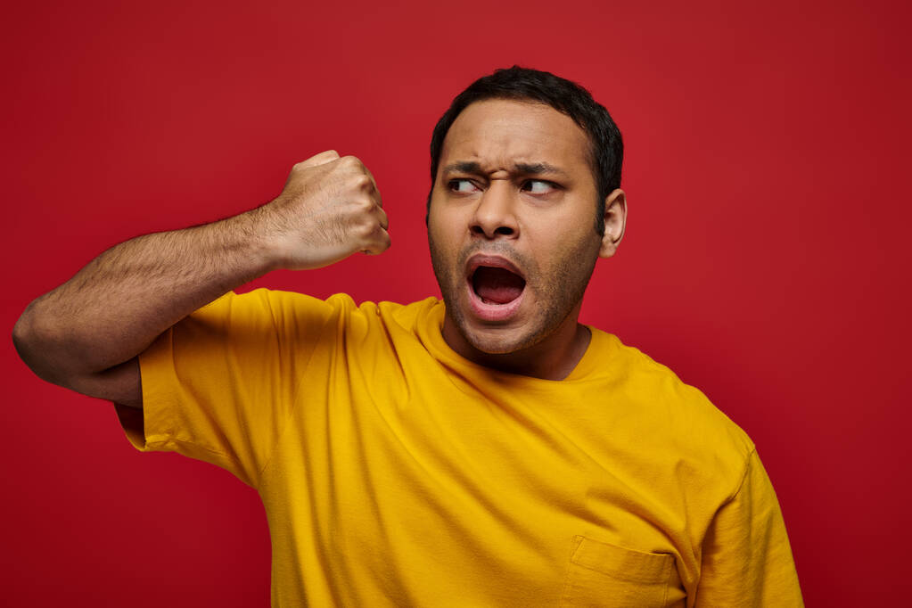 expresión de la cara, hombre indio emocional en camiseta amarilla golpeándose en la cara en el fondo rojo - Foto, imagen