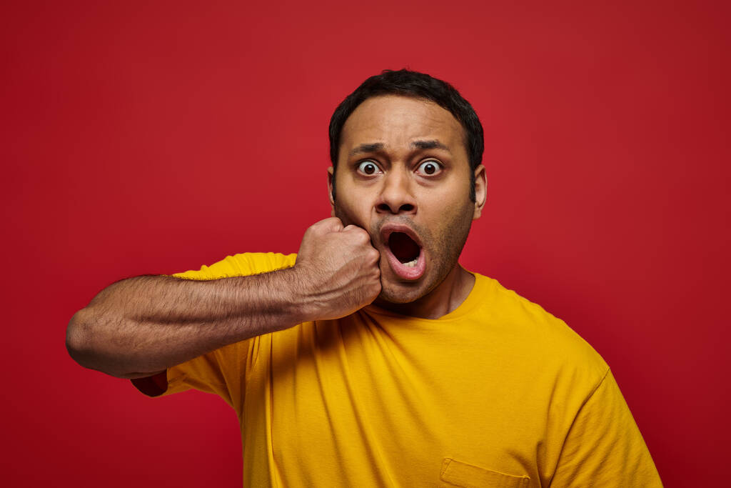 expresión de la cara, impactado hombre indio en camiseta amarilla golpeándose en la cara en el fondo rojo - Foto, imagen