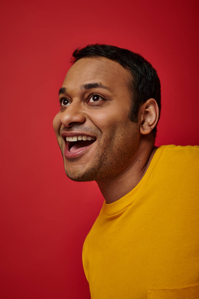 face expression, amazed indian man in yellow t-shirt laughing on red background, open mouth - Photo, Image