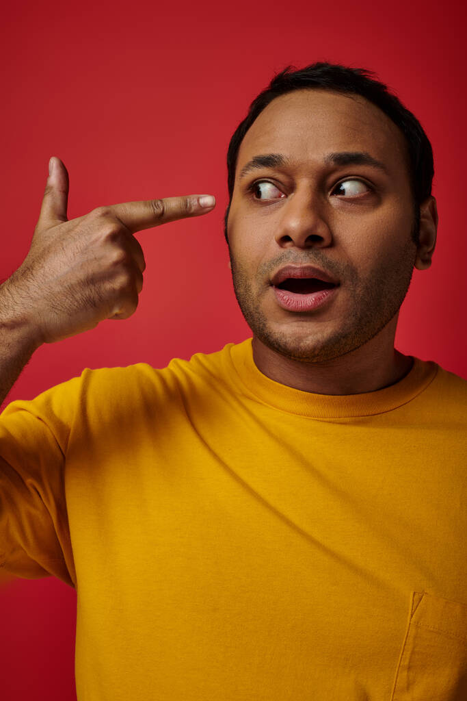 shocked indian man in yellow t-shirt pointing with finger at head on red background in studio - Photo, Image