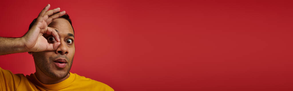 erstaunt indischer Mann in gelbem T-Shirt mit Okay-Zeichen, Hand nahe Gesicht Hintergrund im Studio, Banner - Foto, Bild