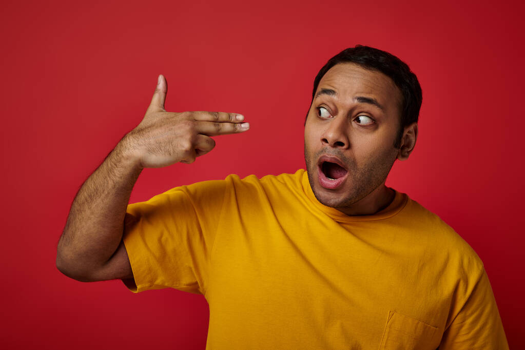 schockierter indischer Mann in gelbem T-Shirt gestikuliert auf rotem Hintergrund und zeigt mit den Fingern in Kopfnähe - Foto, Bild