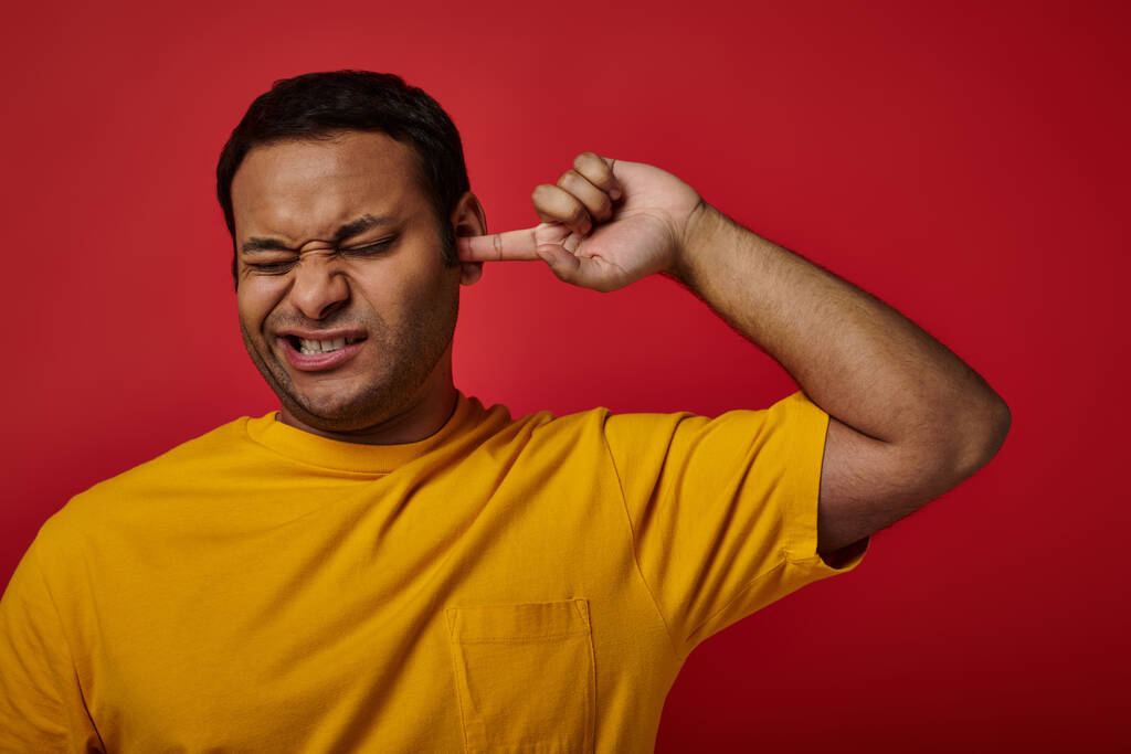 uomo indiano in t-shirt gialla che chiude gli occhi e si raccoglie l'orecchio con un dito su sfondo rosso, smorfia - Foto, immagini
