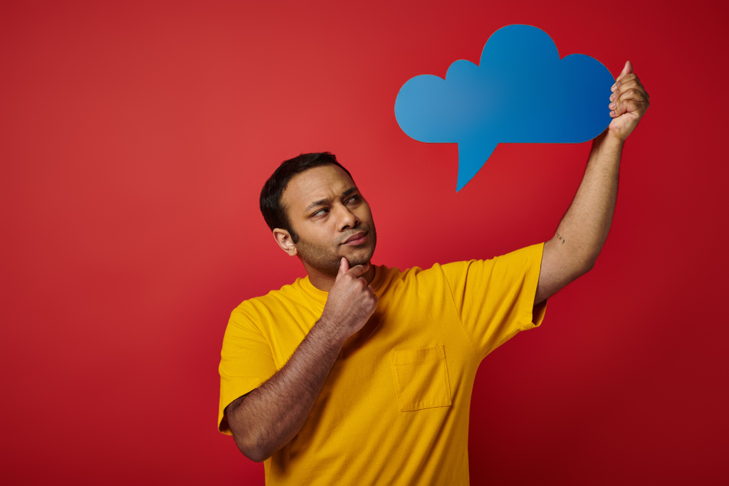 thoughtful indian man in yellow t-shirt holding blank thought bubble on red backdrop, pensive face - Photo, Image
