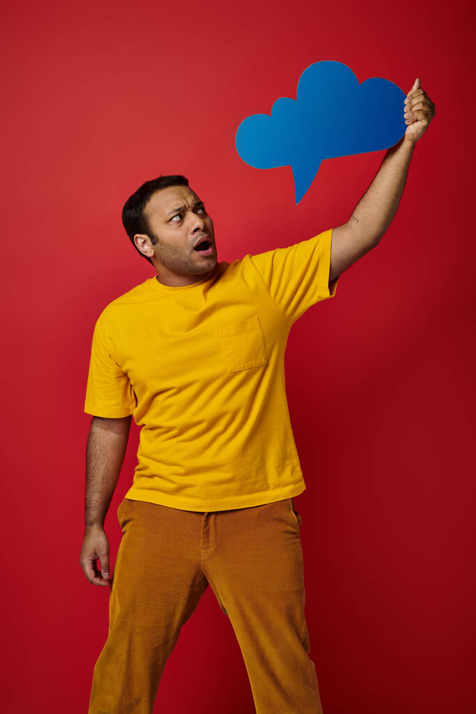 σοκαρισμένος ινδός άνδρας σε κίτρινο t-shirt κρατώντας κενό φούσκα σκέψης στο κόκκινο φόντο, κατάπληκτος πρόσωπο - Φωτογραφία, εικόνα